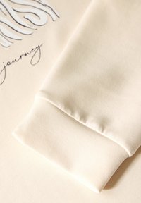 Svetle krémová mikina z textilie s hladkým povrchem; má ohnutý manžet a potištěný design s vlnitými liniemi a kurzívním textem.