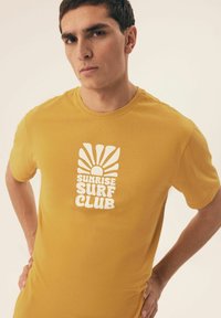 Żółta koszulka z krótkim rękawem z białym nadrukiem graficznym przedstawiającym słońce oraz tekst "SUNRISE SURF CLUB". Wykonana z miękkiego materiału o dopasowanym fasonie.