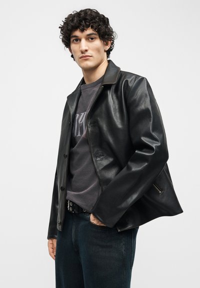 Νεαρός με σγουρά μαλλιά που φοράει μαύρο δερμάτινο μπουφάν πάνω από σκούρο μπλουζάκι με σχέδιο και σκούρο τζιν, στέκεται μπροστά σε μια απλή, φωτεινή φόντο.