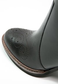 Bottines en cuir noir avec une forme incurvée, présentant des motifs en spirale perforés décoratifs sur le bout et un détail de semelle brune contrastante.
