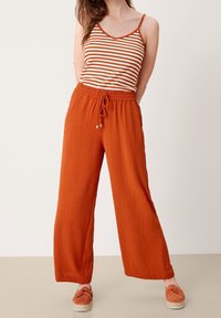 Červenooranžové, texturované široké kalhoty s elastickým pasem a stahovací šňůrkou. Nosič má na sobě pruhovaný tílko a je obutý v espadrilkách.