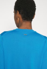 Blå tröja med texturerad stickad design, med rund halsringning och en liten silverlogotyp nära kragen baktill.