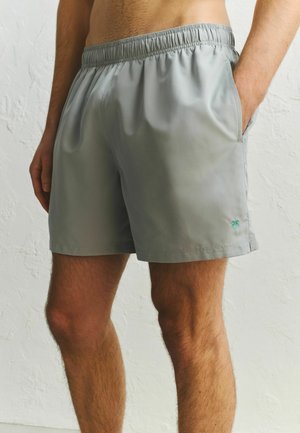 Férfi világosszürke fürdőnadrágban, rugalmas derékpánttal és kis pálmafás logóval a bal oldali zsebén.