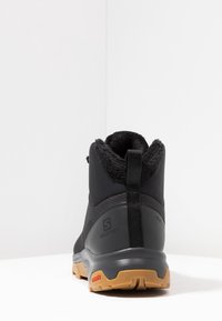 Bota deportiva negra con forro de felpa, suela de goma y parte superior texturizada. Presenta un asa de tirón y un logotipo discreto en el costado.