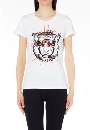 Femme portant un t-shirt blanc avec un motif abstrait coloré de visage de tigre, associé à un pantalon noir, debout sur fond blanc.
