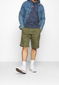 Jeansjacka över marinblå hoodie, olivgröna shorts och vita högskurna sneakers. Funktioner inkluderar framfickor och diskreta metalldetaljer.