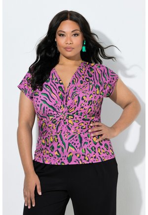 Kobieta o długich ciemnych włosach, nosząca zielone kolczyki, różowy skręcany top w panterkę oraz czarne spodnie, pozuje z jedną ręką na biodrze.