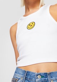 Bílý proužkovaný crop top s kulatou žlutou nášivkou s usměvavou tváří, na které je nápis "NY", a malým červeným detailem, v kombinaci s světle modrými denimovými šortkami.