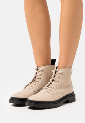 Bottes cheville en daim beige avec semelle en caoutchouc noire, bout rond et œillets métalliques. Comprennent un laçage et un petit passant à l'arrière.