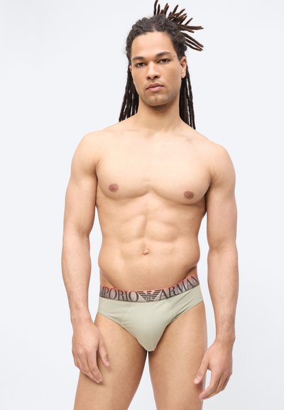 Μυώδης άνδρας με κοτσίδες, φορώντας ανοιχτοπράσινο εσώρουχο Emporio Armani, στέκεται μπροστά σε λευκό απλό φόντο.