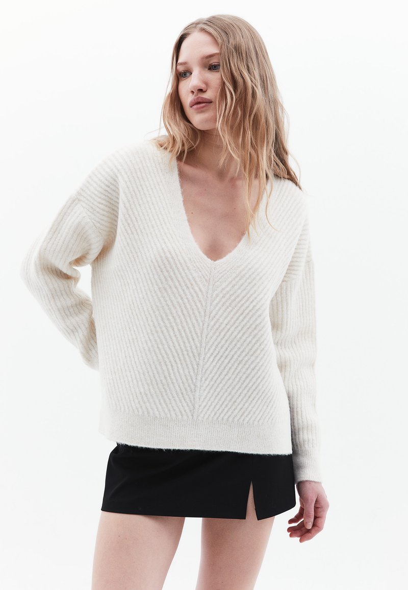 Pullover bianco a coste con scollo a V profondo, maniche lunghe, tessuto testurizzato, abbinato a una mini gonna nera con uno spacco laterale.