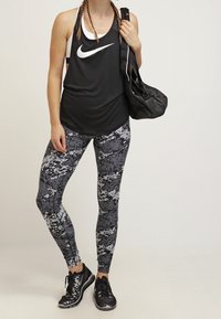 Černý sportovní tílko s bílým logem Nike, šedé legíny se vzorem a černé tenisky. Nese černou tašku.