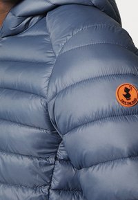 Blue puffer jacket with horizontal quilted sections. Features a small orange circular logo on the upper arm. Smooth, shiny fabric texture.