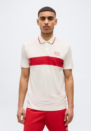 Jeune homme portant un polo blanc avec une bande horizontale rouge et un short rouge, debout devant un fond clair uni.