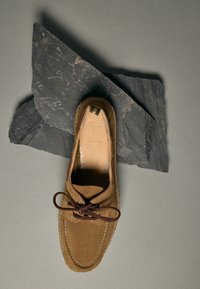 Mocassin en daim marron avec un lacet en cuir noué en nœud, posé sur deux dalles en ardoise foncées sur un fond gris clair.