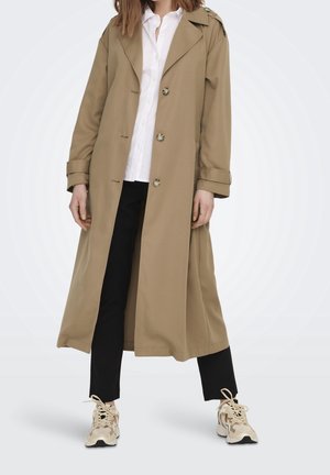 Person, die einen langen beigen Trenchcoat über einem weißen Hemd, schwarzen Hosen und beigen Sneakers trägt und vor einem schlichten hellen Hintergrund steht.