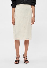 Saia midi branca bordada com padrões florais, feita de tecido leve. Usada com sandálias pretas de tiras.