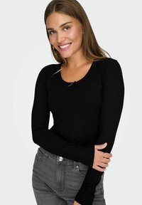 Zwarte ribbeltrui met lange mouwen en een ronde halslijn, versierd met een klein strikje in het midden. G gedragen met grijze hoge taille jeans.
