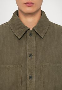 Camicia in velluto a coste verde oliva con colletto, due tasche anteriori e bottoni neri, caratterizzata da una texture a coste e un design classico.