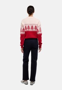 Pull en tricot rouge et crème avec des motifs d'arbre festifs, associé à un jean foncé et des chaussures argentées. Accent sur le design géométrique et la texture.