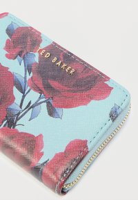 Portofel albastru cu fermoar, decorat cu imprimeu de trandafir roșu și logo auriu "Ted Baker" pe o suprafață texturată.