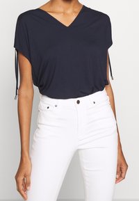 Námořnický top s výstřihem do V a krátkými rukávy, s nastavovacími bočními šňůrkami, kombinovaný s bílými džíny s vysokým pasem. Měkká tkanina, volný střih, minimalistický design.