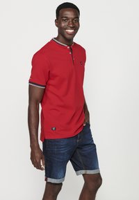 Tricou polo scurt roșu, cu un finisaj texturat, două butoane la guler și un guler cu dungi contrastante. Asortat cu pantaloni scurți din denim.