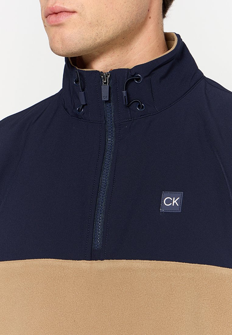 Gros plan sur une personne portant un manteau zippé bleu marine et beige avec des cordons réglables au col et un patch logo "CK".