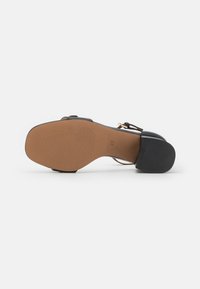 Sandal con tacco nero, dotato di una cinghia liscia, suola testurizzata e suola color tan. Presenta una cinghia sottile alla caviglia e un design a punta tondeggiante.