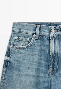 Niebieskie dżinsy z denimu w talii, z pętlami na pasek, metalowym guzikiem, widocznymi przednimi kieszeniami oraz kieszonką na bilon na białym tle.