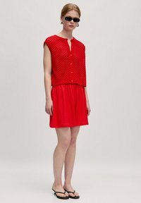 Rotes gestricktes Oberteil mit durchbrochenem Design und kurzen Ärmeln, kombiniert mit einem roten, gerafften Rock. Schwarze Flip-Flops runden das Outfit ab.