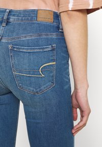 Blå denimjeans med en figursydd design, med en läderpatch märkt "American Eagle," och ett utmärkande guldstickningsmönster på bakfickan.