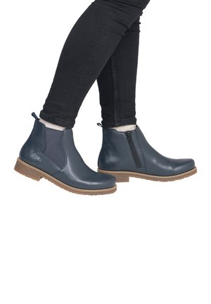 Person wearing dark blue leather ankle boots with brown soles, black pants, and white socks, shown from the side against white background.