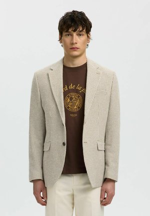 Ung mann iført beige hundetannt blazere, brun grafisk t-skjorte og kremfargede bukser, står foran en ensfarget lys bakgrunn.