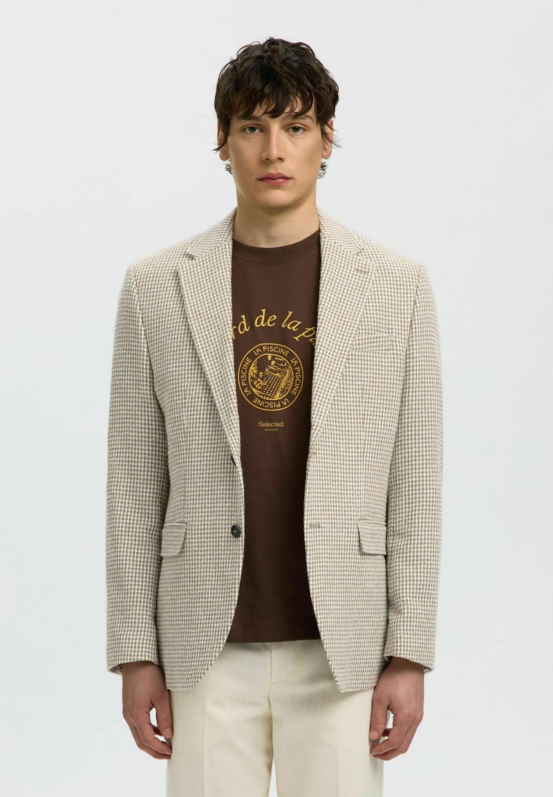 Joven con blazer beig de pata de gallo, camiseta marrón con estampado gráfico y pantalones crema, de pie frente a un fondo claro y liso.