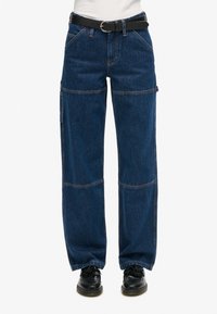 Pantalones cargo de mezclilla azul oscuro con piernas anchas, que presentan múltiples bolsillos, costuras contrastantes y un cinturón negro. Material texturizado y duradero.