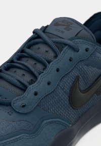 Zila sporta apavi ar acu tīklu un zamšu materiālu, plakanas auklas un noapaļotu purngalu. Iezīmē melnu Nike logo un nelielus teksturētus akcentus.