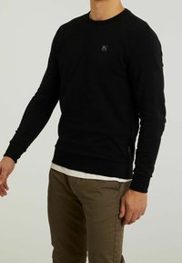 Homme portant une chemise noire à manches longues avec un petit logo sur la poitrine et un pantalon marron, debout devant un fond clair uni.