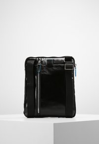 Bolso de cuero negro con un bolsillo frontal con cremallera, correas ajustables y un pequeño acento azul cerca de la parte superior. Textura suave y diseño minimalista.