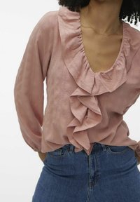 Blouse rose clair avec encolure à volants et manches longues, en tissu texturé et décolleté en V profond. Associée à un jean en denim bleu.