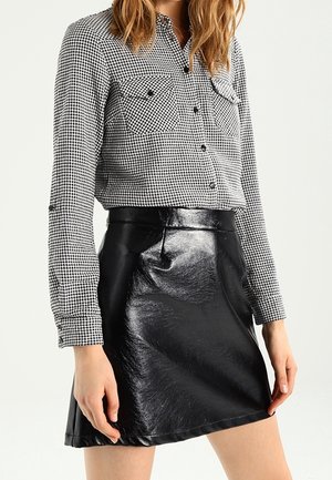 Frau trägt ein schwarz-weißes Hahnentritt-Knopfhemd, das in einen glänzenden schwarzen Leder-Minirock gesteckt ist.