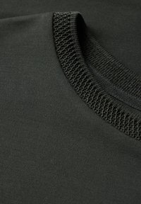 Tmavě zelená bavlněná látka s žebrovým výstřihem. Textura je hladká a zvýrazňuje pletený vzor kolem límce.