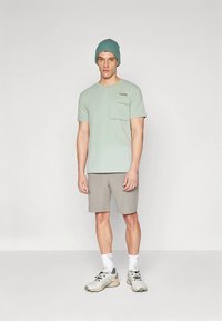 Mann som står i lysegrønn kortermet skjorte med lommer, grå shorts, grønn lue, hvite sokker og grå joggesko på ensfarget bakgrunn.