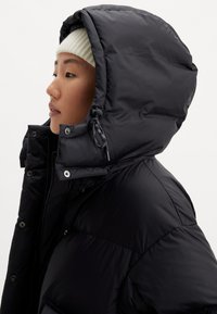 Person wearing a black hooded puffer jacket and white knit beanie, facing left, against a plain light background.