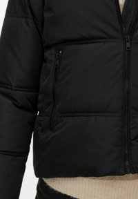 Kort svart pufferjakke med glatt tekstur, med sidelommer med glidelås og horisontal sømdetalj langs ermene.