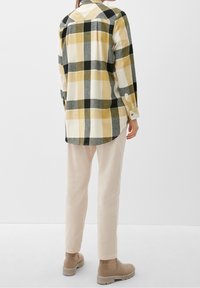 Camicia di flanella con un motivo a quadri giallo, nero e grigio. Maniche lunghe, polsini con bottone. Abbinata a pantaloni chiari e stivali beige.