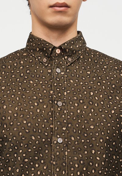 Άνδρας που φοράει καφέ πουκάμισο με κουμπιά και μαύρο-μπεζ λεοπάρ μοτίβο, εμφανίζοντας το γιακά και την περιοχή του άνω στήθους.