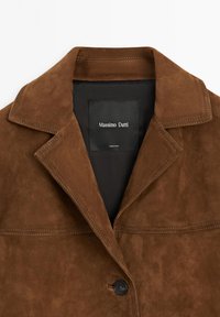 Brązowa kurtka zamszowa z wyłogami, pojedynczym zapięciem na guzik i wewnętrzną metką. Teksturowany materiał z widocznym przeszyciem.