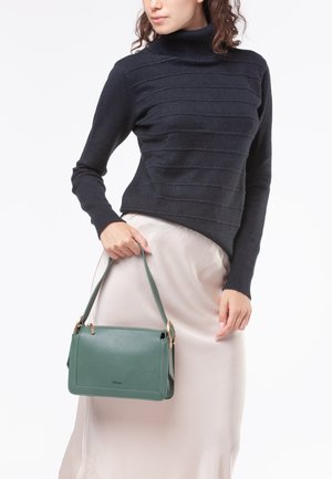 Borsa a tracolla in pelle verde con un design strutturato, accessori in oro e una tracolla rimovibile, tenuta da una persona che indossa un top scuro e una gonna chiara.