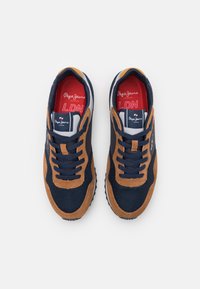 Marina och tan mockaskor med textilöverdel, snörning fram, texturerad sula och märkta innersulor med röd och vit text.